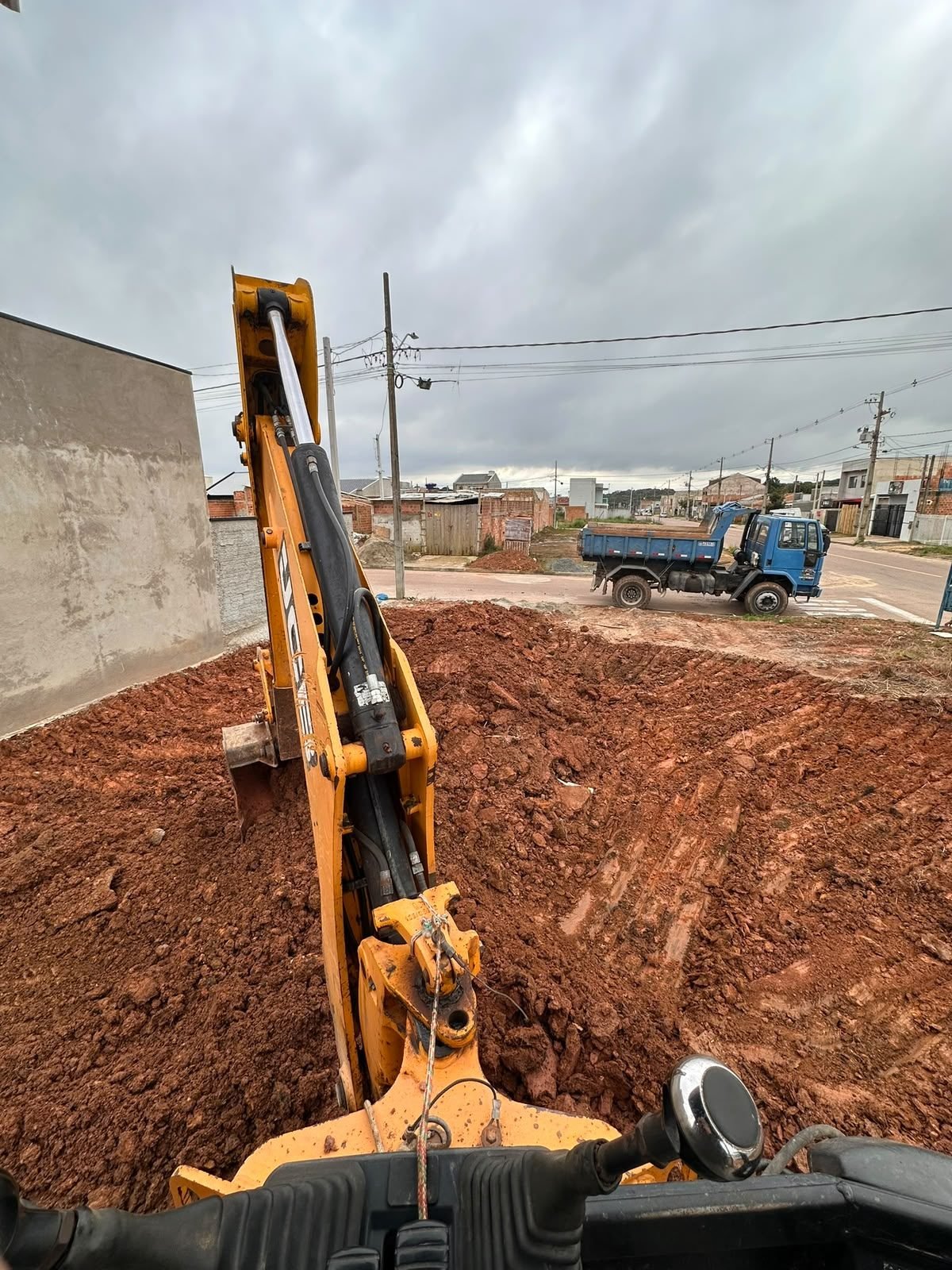 Retroescavadeira escavando vala em terreno residencial com caminhão ao fundo